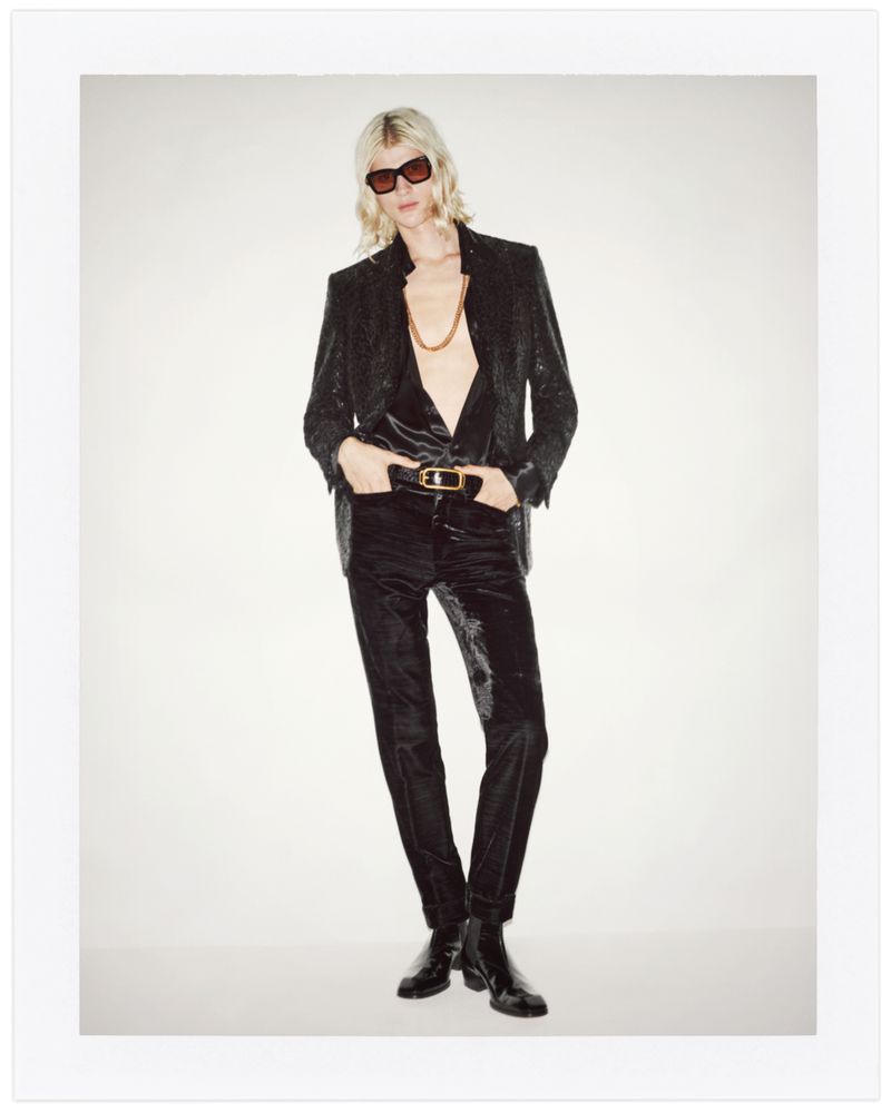 Full-length shot of a slender adult woman with wavy blond hair in a black sparkly blazer and pants, posing in front of a white background.