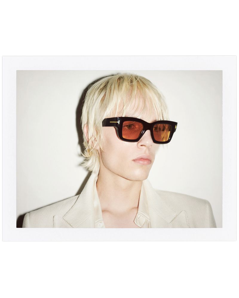 Headshot of person with blonde hair and orange sunglasses in a cream-colored jacket against a white background in portrait orientation.