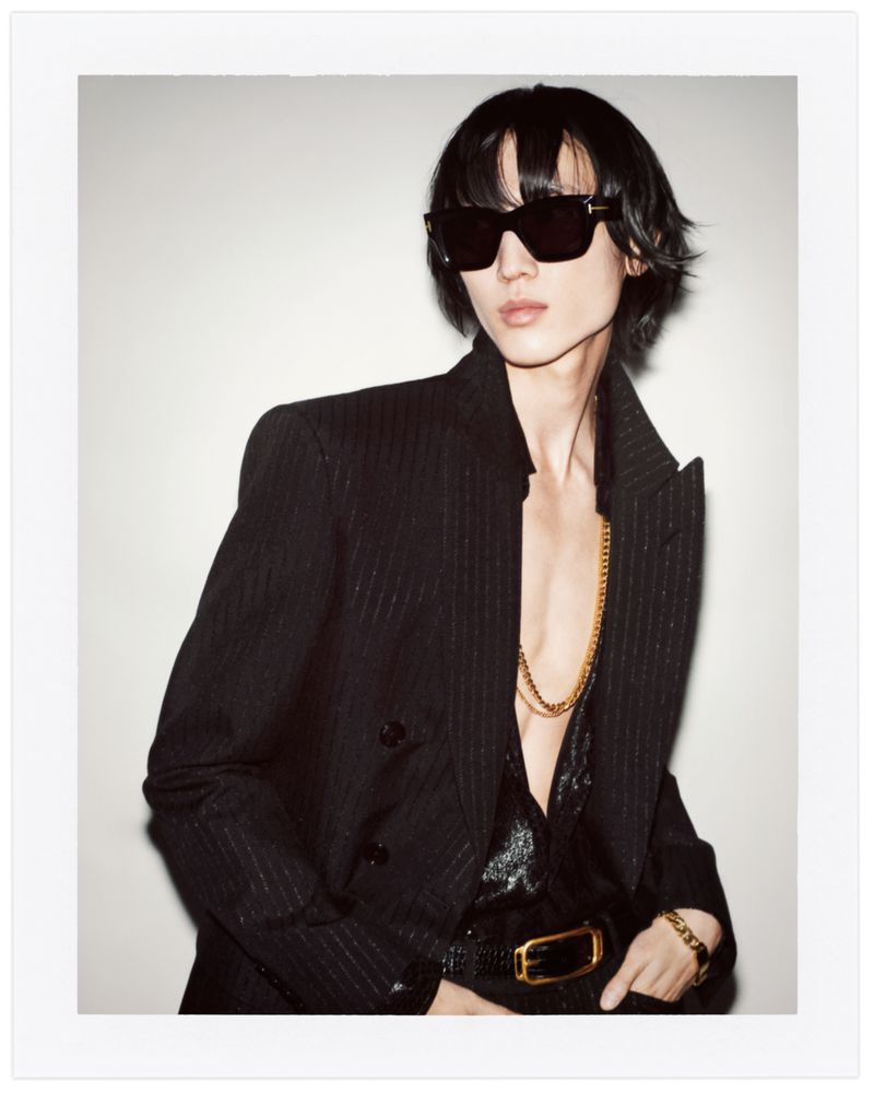 Adult male poses indoors wearing a dark pinstripe suit, black sunglasses, and gold jewelry against a neutral background in a fashion shot.