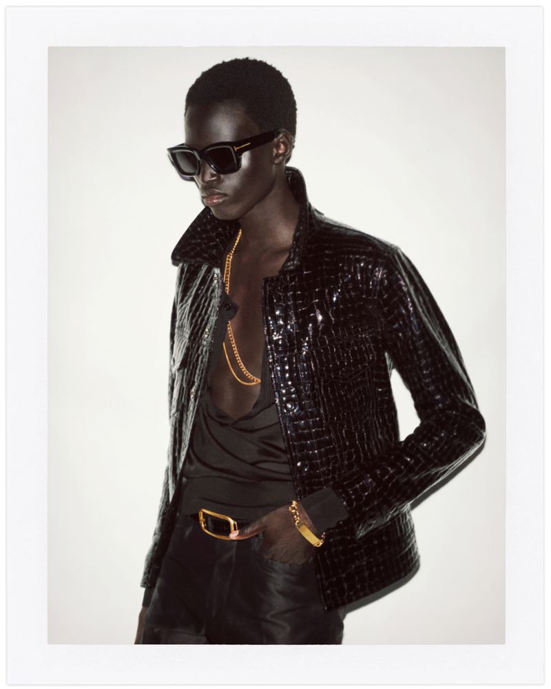A person poses in a black jacket and sunglasses against a white background. The jacket has a reptile skin pattern and gold jewelry accents.