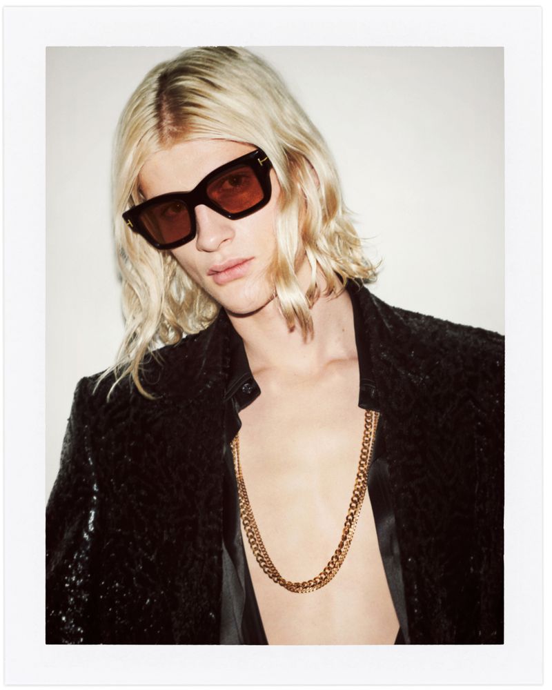 Portrait of a blond male model wearing a black jacket, sunglasses, and gold chain, posing against a bright white backdrop.