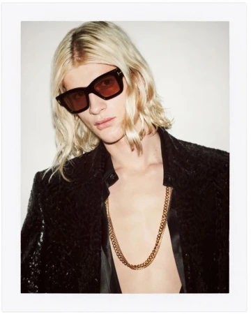 Portrait of a blond male model wearing a black jacket, sunglasses, and gold chain, posing against a bright white backdrop.
