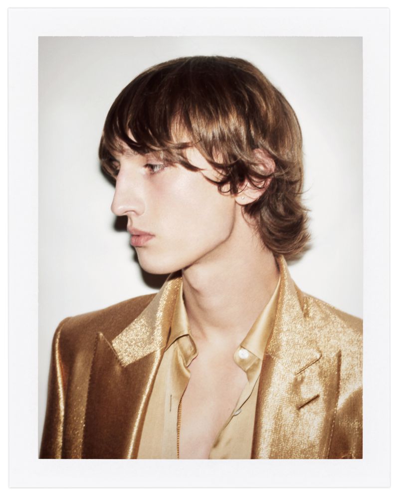 Profile shot of a young adult male with shoulder-length brown hair, wearing a shiny gold blazer and collared shirt against a white backdrop.