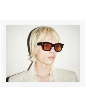 Model wearing dark sunglasses with tinted lenses and a light cream blazer against a plain white studio backdrop.