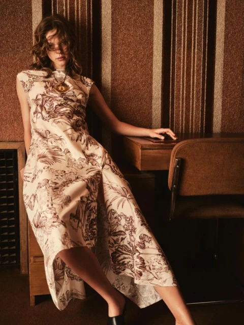 A woman poses in a patterned dress next to a desk in an indoor setting with brown striped wallpaper, fashion shoot.