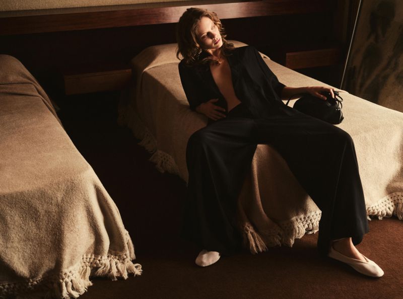 Adult woman lounges on a bed in an open black robe, wide-legged pants, and ballet flats, holding a small black purse near another bed.