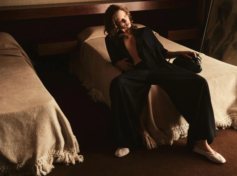 Adult woman lounges on a bed in an open black robe, wide-legged pants, and ballet flats, holding a small black purse near another bed.