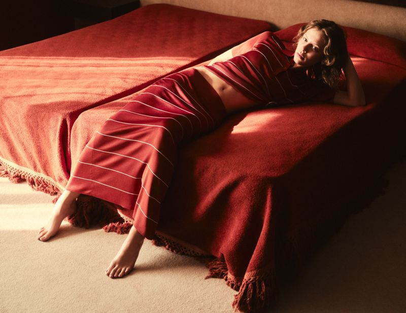 Adult female lying on a red bed with white striped clothing indoors during daytime. The background is blurred, and the light is soft.