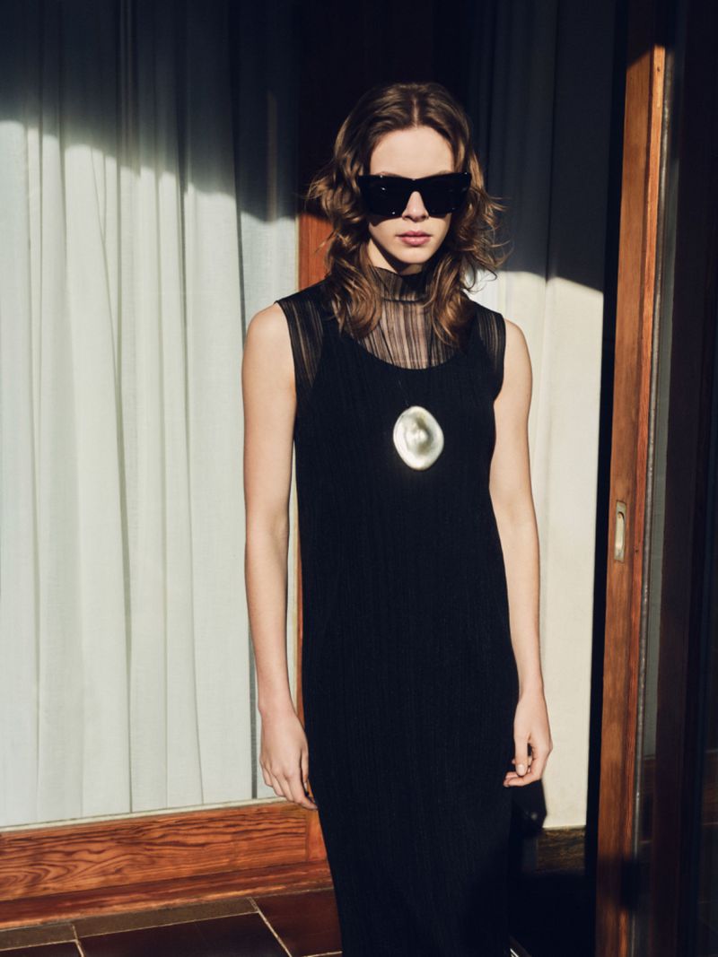 A stylish adult female model poses indoors near a window, wearing a black dress, sunglasses, and a large, light-colored pendant necklace.
