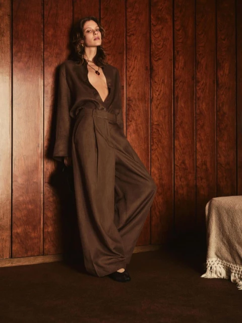 Adult female fashion model poses in an open brown top and loose brown pants in an indoor studio with wood wall paneling.