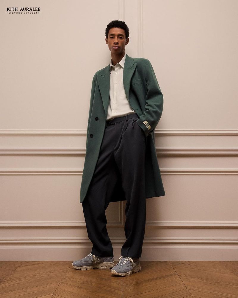 An adult male stands in a photo studio, wearing a long green coat, dress pants, and sneakers; he is leaning against a cream-colored wall.