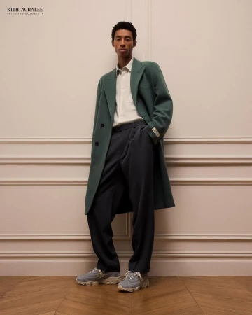 An adult male stands in a photo studio, wearing a long green coat, dress pants, and sneakers; he is leaning against a cream-colored wall.