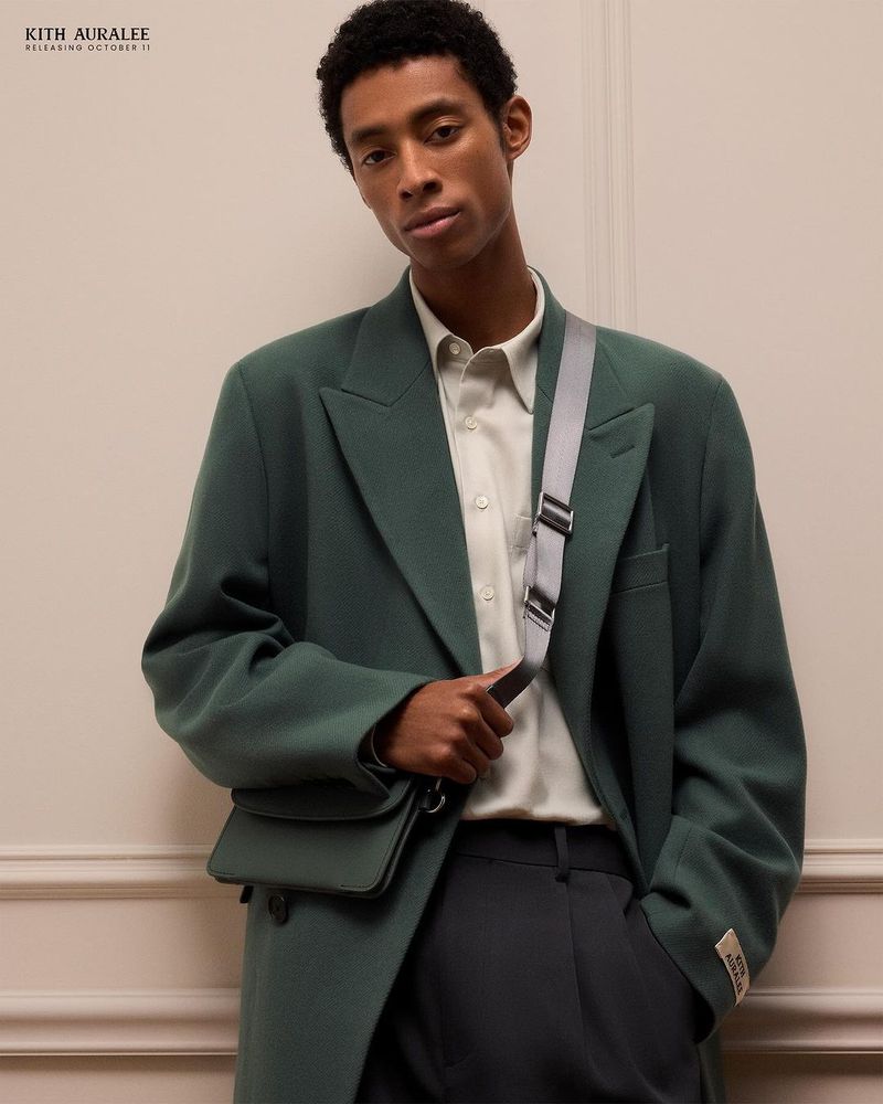 A young adult man wearing a green jacket and shoulder bag looks at the camera against a neutral wall backdrop indoors.