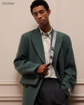 A young adult man wearing a green jacket and shoulder bag looks at the camera against a neutral wall backdrop indoors.