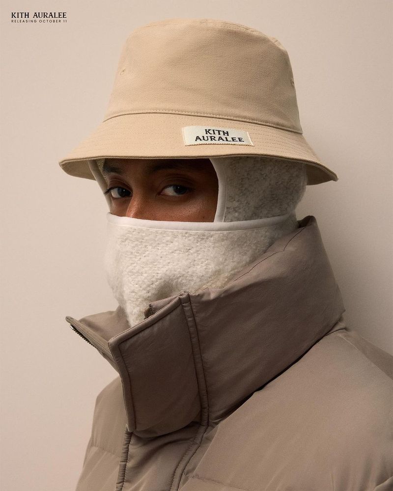 Person wearing a beige bucket hat, a white balaclava, and a beige puffer jacket poses against an off-white seamless backdrop.