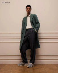 Man wearing a green wool coat, white button-down shirt, dark trousers, and sneakers standing against a classic paneled wall.