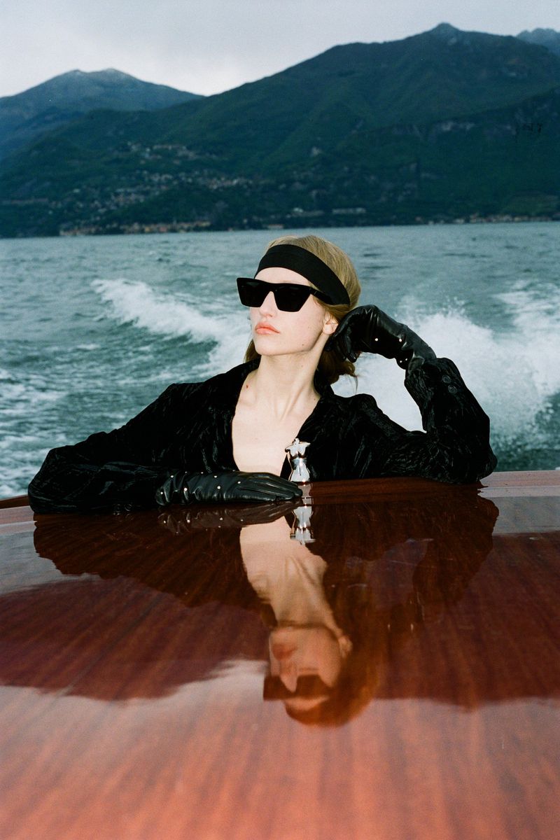 Elegant woman in sunglasses and velvet jacket on a boat, with mountains and water in the background, creating a luxurious and stylish scene.