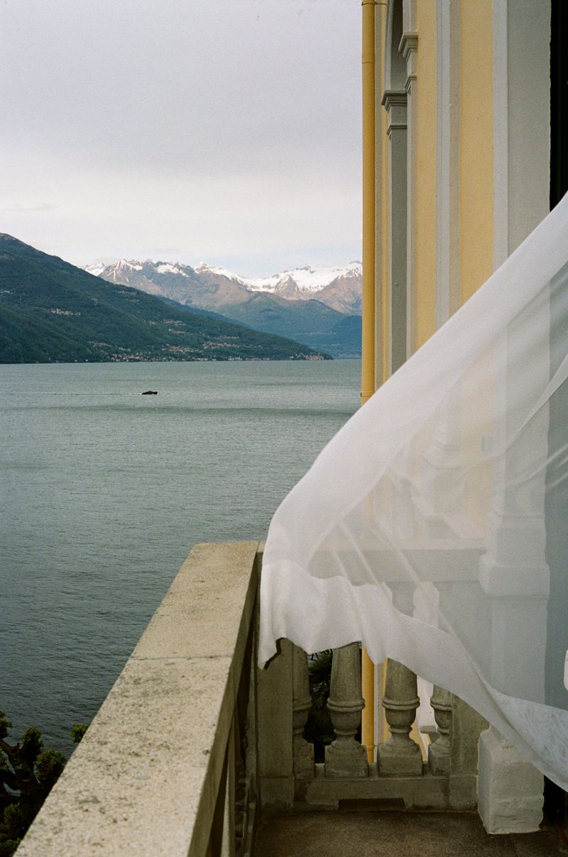 Scenic view from a balcony overlooking a lake and snow-capped mountains, with a sheer white curtain partially obscuring the view.