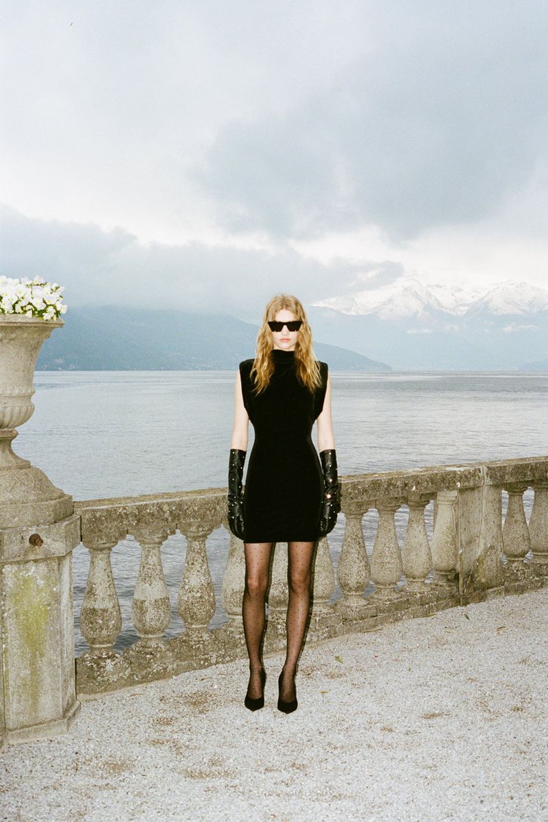 Adult female fashion model poses in a black mini dress, gloves, sunglasses and heels on a stone patio overlooking a lake and mountains.