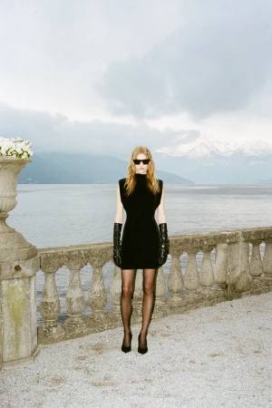 Adult female fashion model poses in a black mini dress, gloves, sunglasses and heels on a stone patio overlooking a lake and mountains.