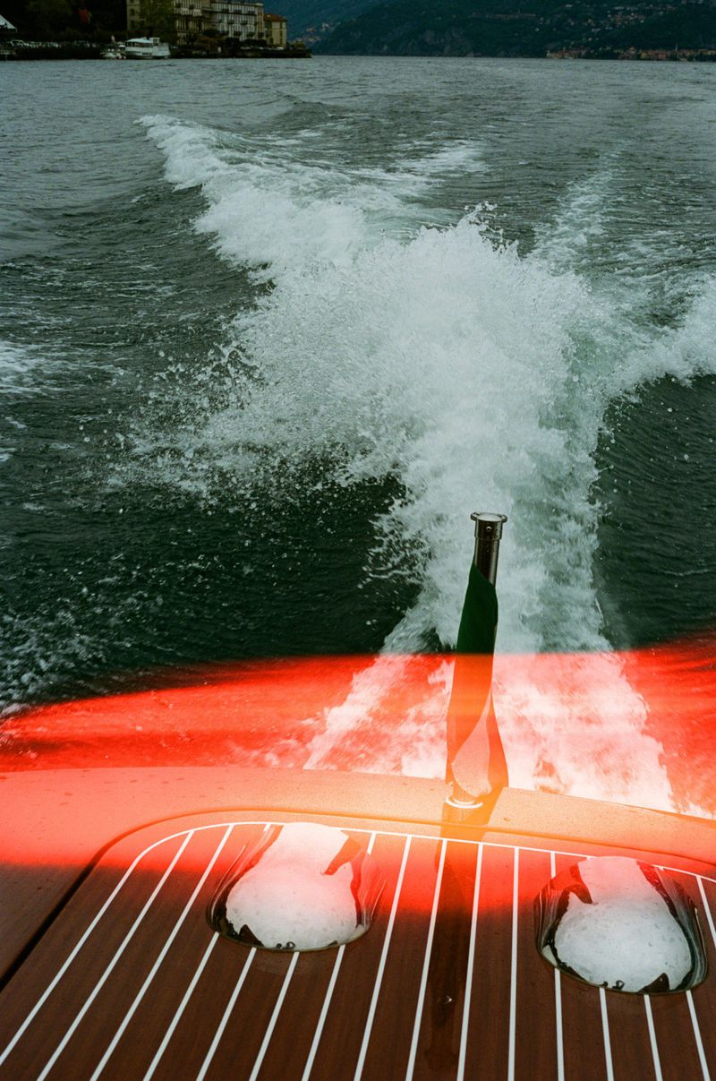 Boat's stern view with brown deck, chrome accents, and Italian flag, creating a wake on the green water of Lake Como.