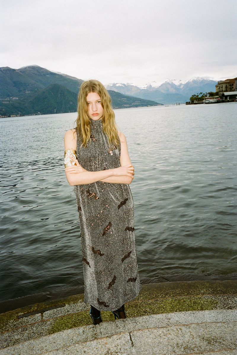 Full-length shot of a fashionable, young adult female model posing in a shiny, sleeveless midi dress by a large lake with a mountain backdrop.
