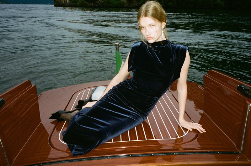 A woman in a dark velvet dress is sitting on the polished wooden deck of a boat, with water and forested shore in the background.