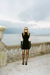 Model in black dress and long leather gloves standing by a stone balustrade with a lake and mountain backdrop under an overcast sky.