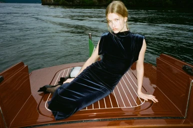 A female model wearing a dark velvet dress lounging on the wooden deck of a boat on open water