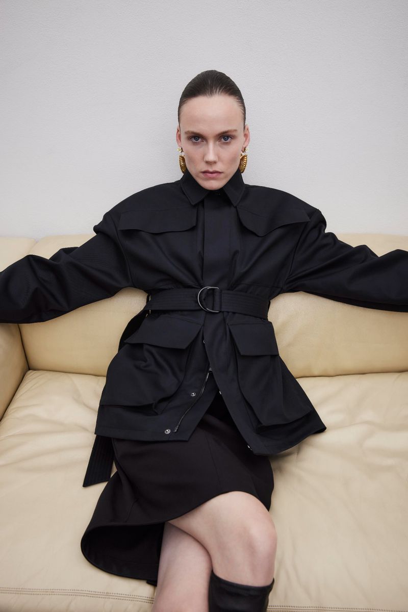 A stylish woman in a black belted jacket and skirt sits on a light yellow couch, accessorized with gold earrings and dark boots.
