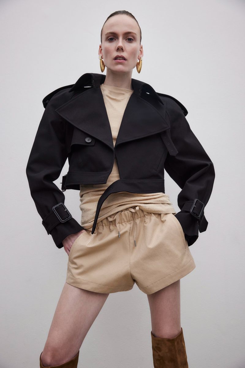 A slender adult woman poses indoors, wearing a black cropped trench coat, beige shorts, and brown boots against a neutral backdrop.