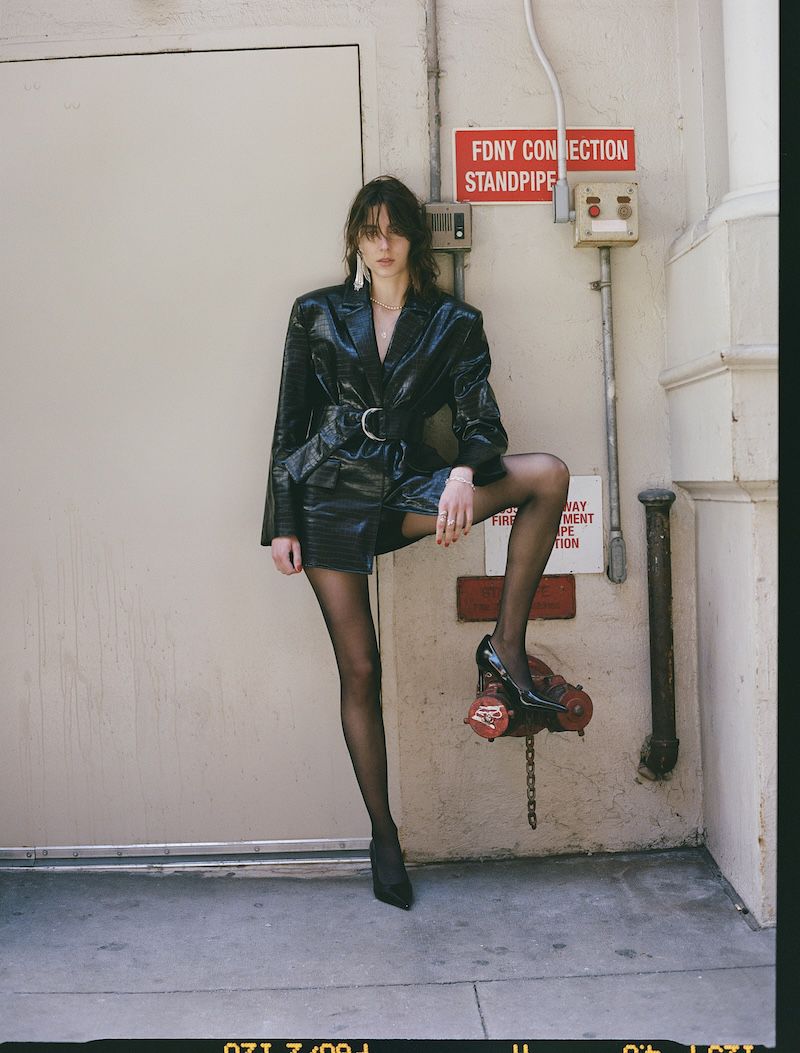 A woman in a shiny black jacket and skirt poses in front of a fire hydrant and the side of a building with an FDNY connection box.