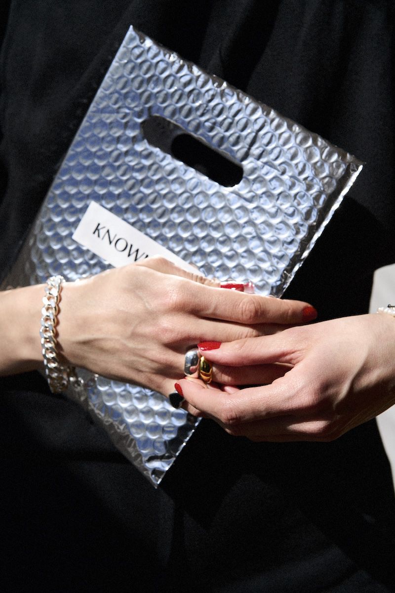 Close-up shot of silver textured bag, adorned with two gold and silver rings held by a hand with red nail polish, with silver bracelet.