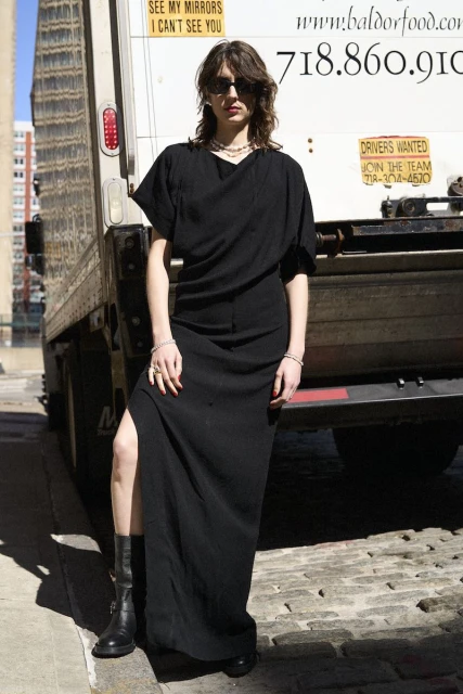 Woman in black maxi dress poses near delivery truck on city street. She is wearing sunglasses and boots in natural daylight.
