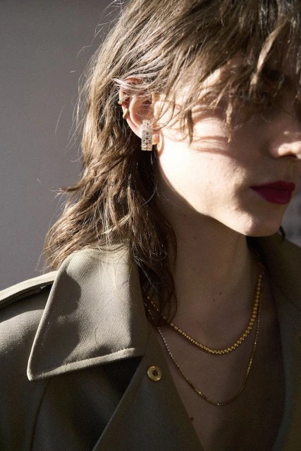 Close-up of a woman with light brown hair wearing square earrings, a green coat, and gold necklaces against a neutral background.