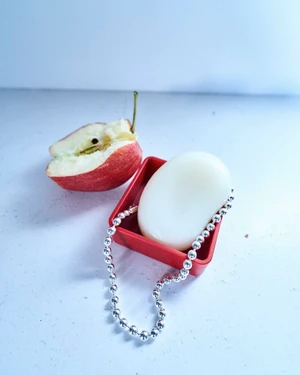 Close-up of a silver bead necklace on a red tray next to a sliced red apple on a pale surface.
