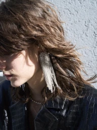 Close-up profile of a model wearing a large crystal fringe earring and textured black jacket against a grey textured background.