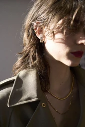 Close-up portrait of a woman wearing gold hoop earrings, layered necklaces, and an olive trench coat in natural sunlight.