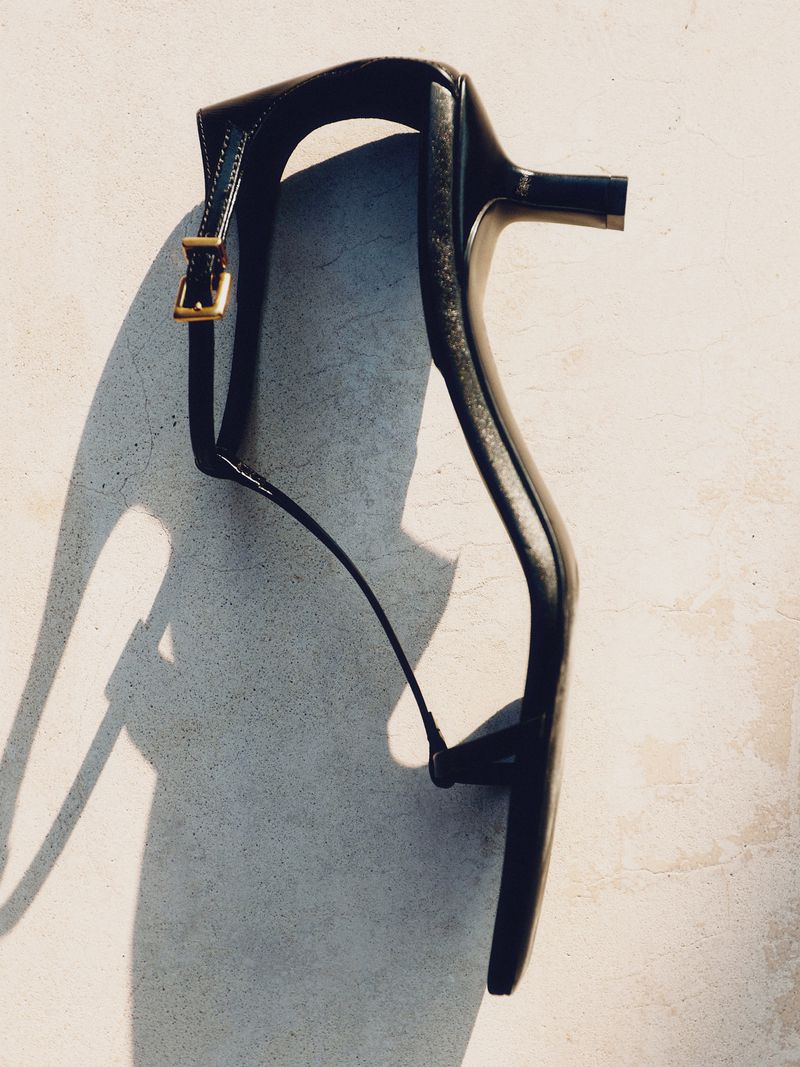 Elevated view of a black, high-heeled sandal with a delicate ankle strap and gold buckle, casting a soft shadow on an off-white surface.