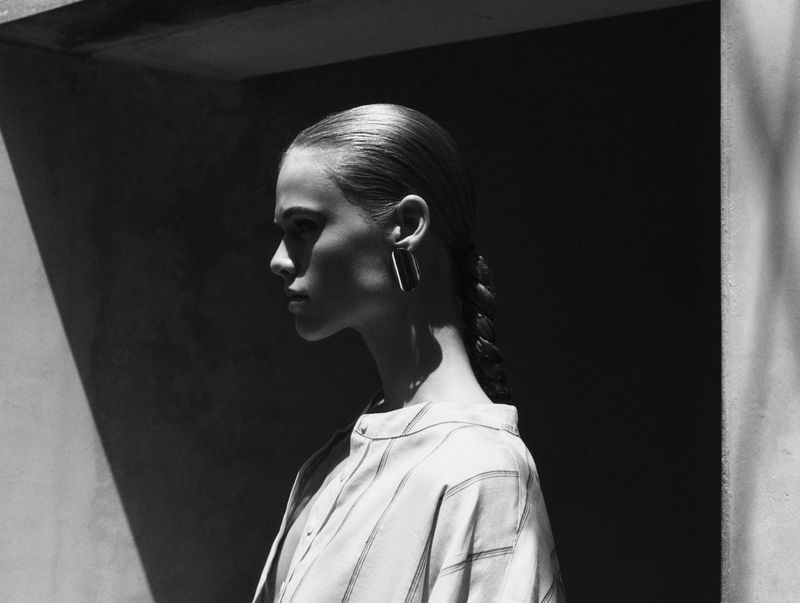 Black and white portrait of a young woman in profile wearing a collared shirt and earrings, standing in a shadowed outdoor space.