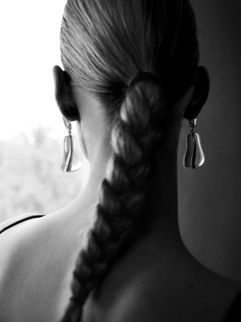 Black and white studio shot of a woman's back and braided hair, wearing dangle earrings, against a light background.