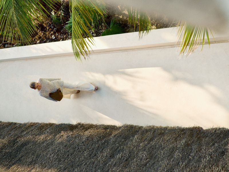 High-angle view of a woman walking on a sunlit path, wearing white linen clothing and carrying a shoulder bag, with palm trees overhead.