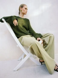 Model seated on a white folding chair wearing a green knit sweater and light green trousers against a neutral wall