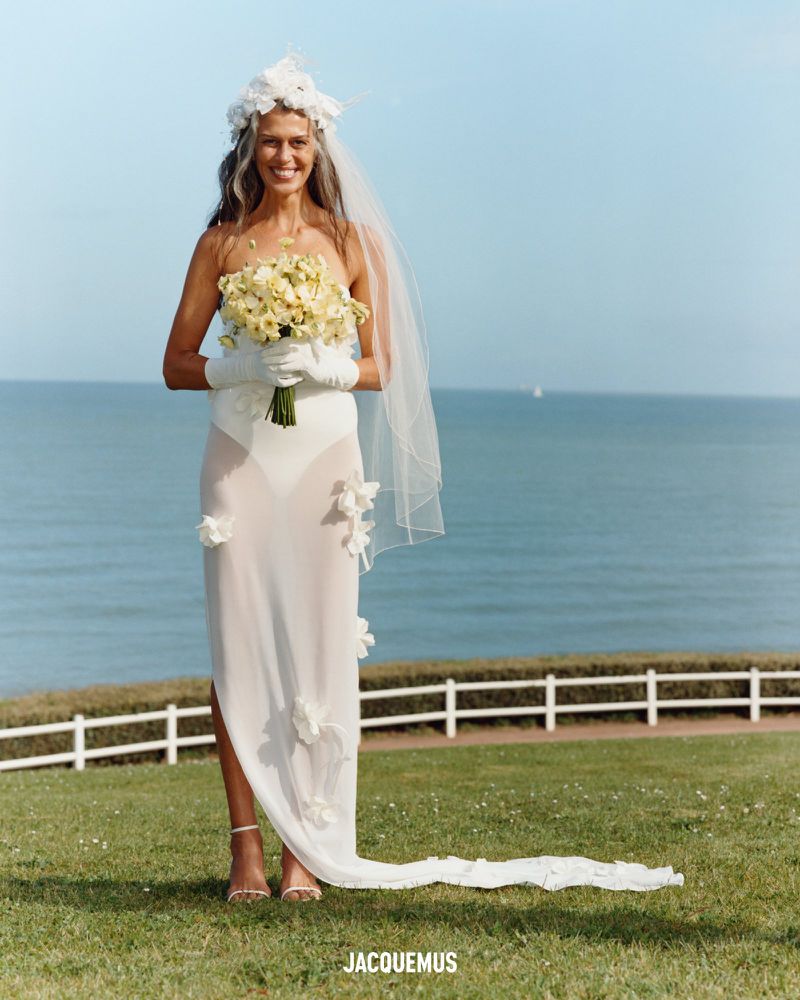 Woman in a white bridal gown holds a bouquet of flowers in front of the ocean on a sunny day, fashion editorial shot, Jacquemus branded.