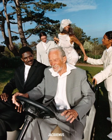 A diverse group of models including an older male in a grey suit, smiling, are seated in a golf cart in a grassy, sunny landscape.