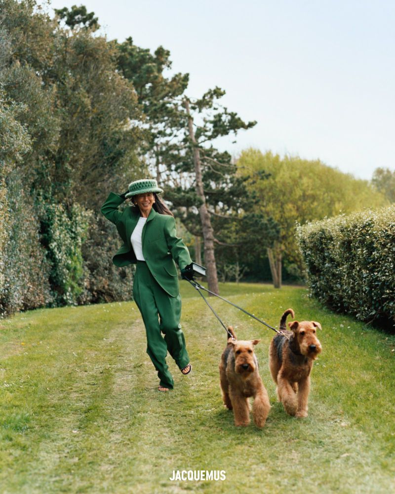 A woman in a green suit walks two Airedale Terrier dogs on a grassy path in a sunlit park, wearing a matching green hat, with JACQUEMUS logo text.