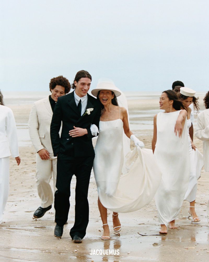 Wedding party on a beach, featuring a bride in a white dress and hat, arm-in-arm with the groom in a black suit and tie.