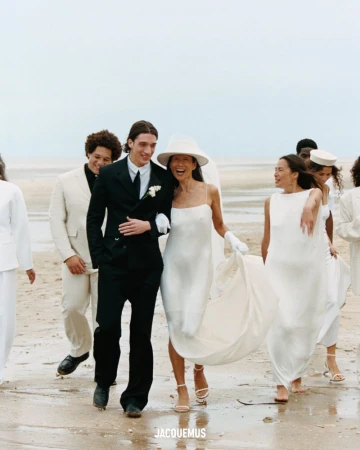 Wedding party on a beach, featuring a bride in a white dress and hat, arm-in-arm with the groom in a black suit and tie.