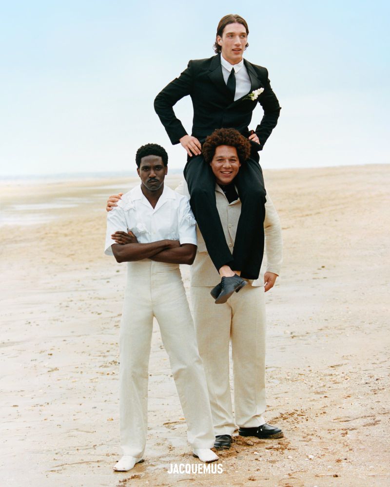 Three fashion models pose on a beach; one is Black with crossed arms, another carries the first one on his shoulders.