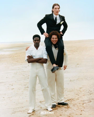 Three fashion models pose on a beach; one is Black with crossed arms, another carries the first one on his shoulders.
